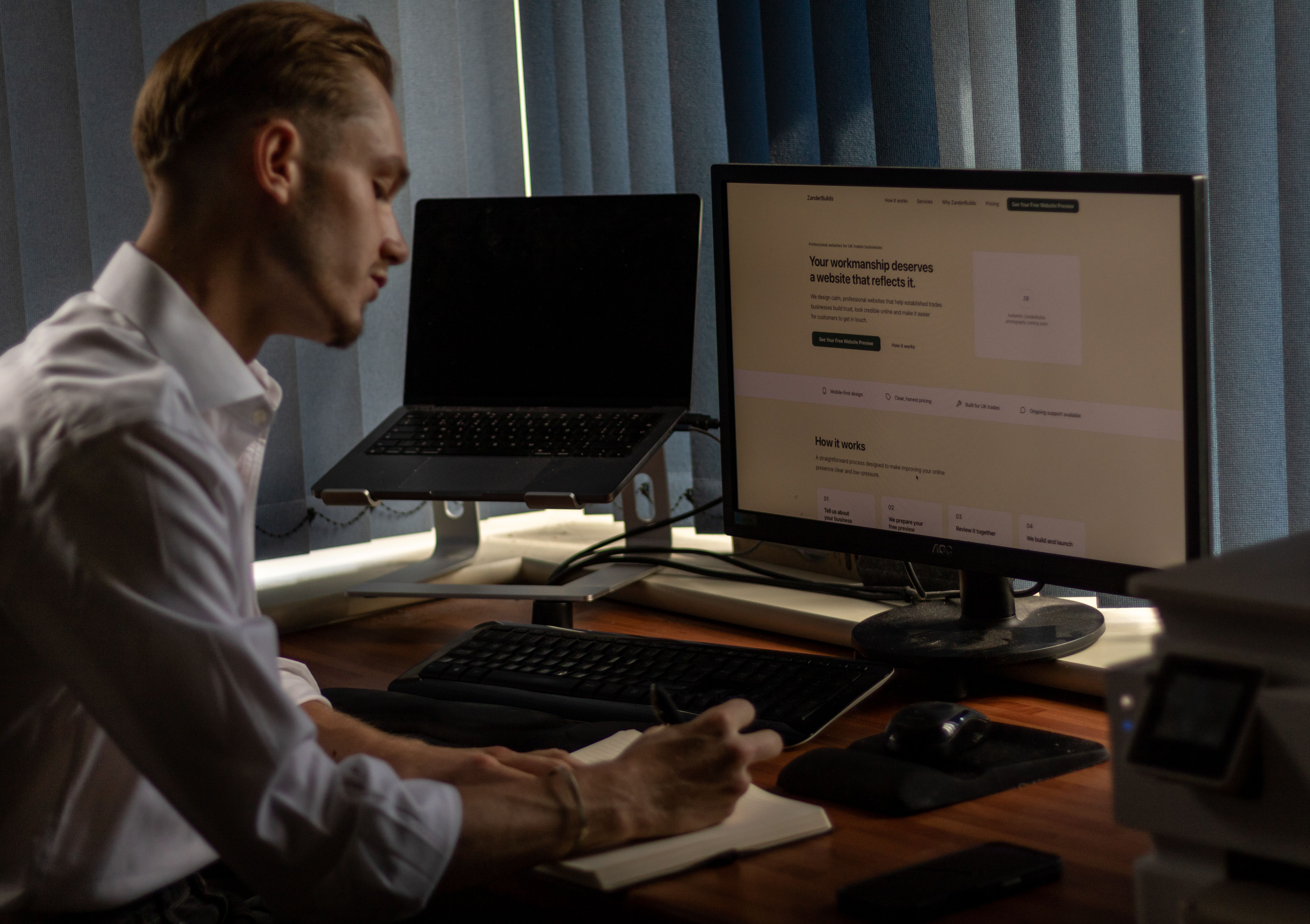 Alex planning in a notebook at his desk, with the BuiltByAL homepage open on the monitor beside him.